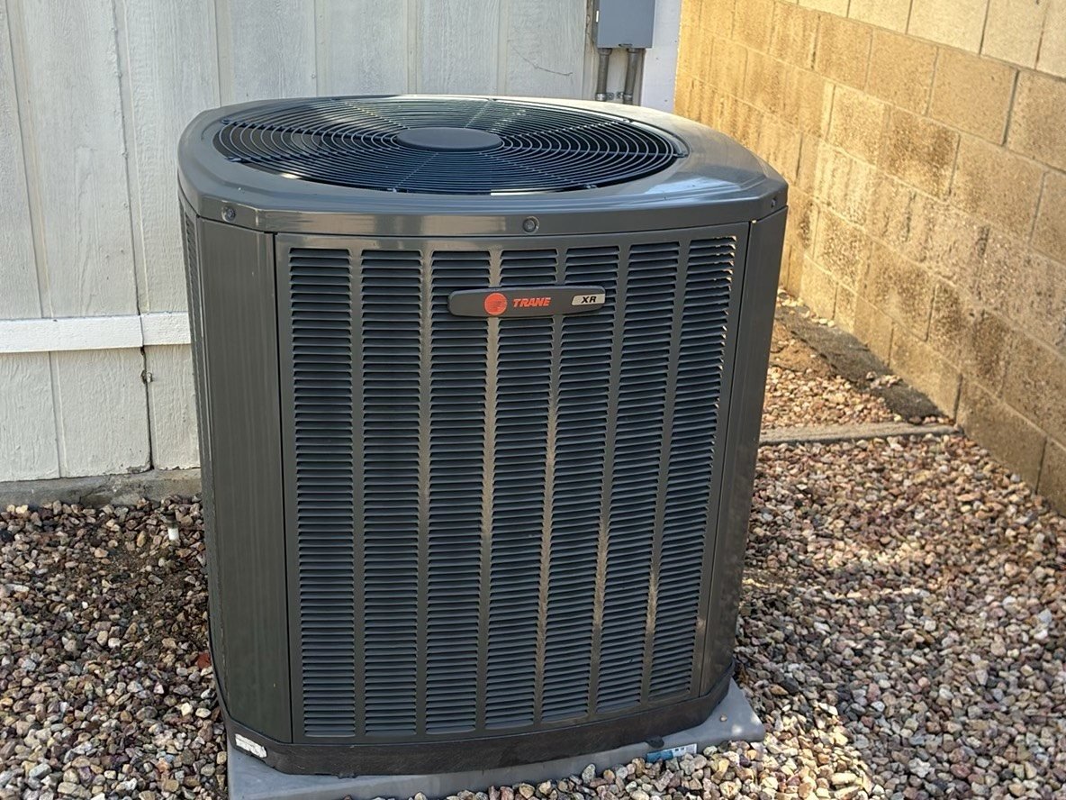 A Trane brand central air conditioning condenser unit sitting on a gravel pad next to a block wall fence, sharp and well-lit