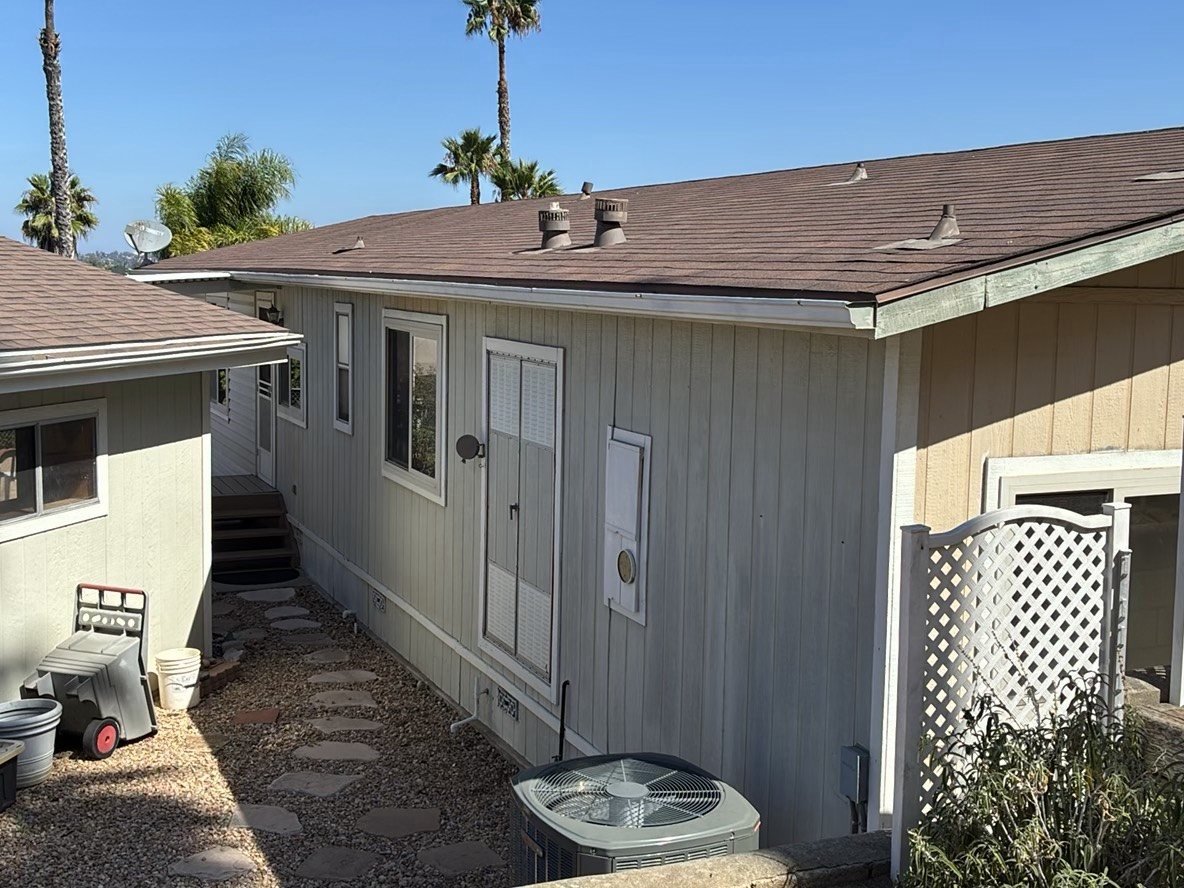 Wide exterior shot of a single-story home with brown asphalt shingle roof, outdoor AC condenser unit, and palm trees in the background under a clear blue sky