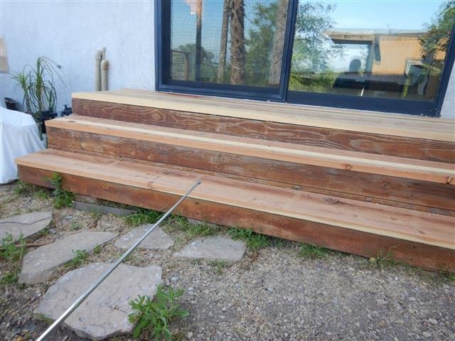 Contemporary home exterior showing warm cedar stair treads leading to a large black-framed sliding glass door with tropical garden and stone steppers