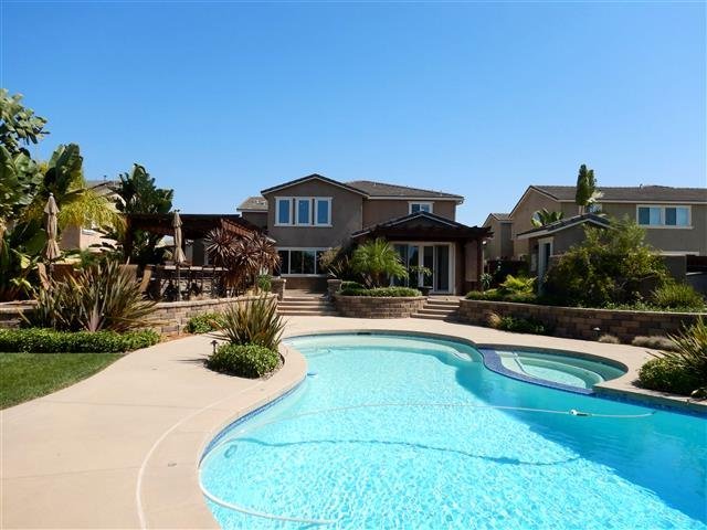Freeform swimming pool with attached spa adjacent to a two-story stucco estate home with mature tropical landscaping under a clear blue sky